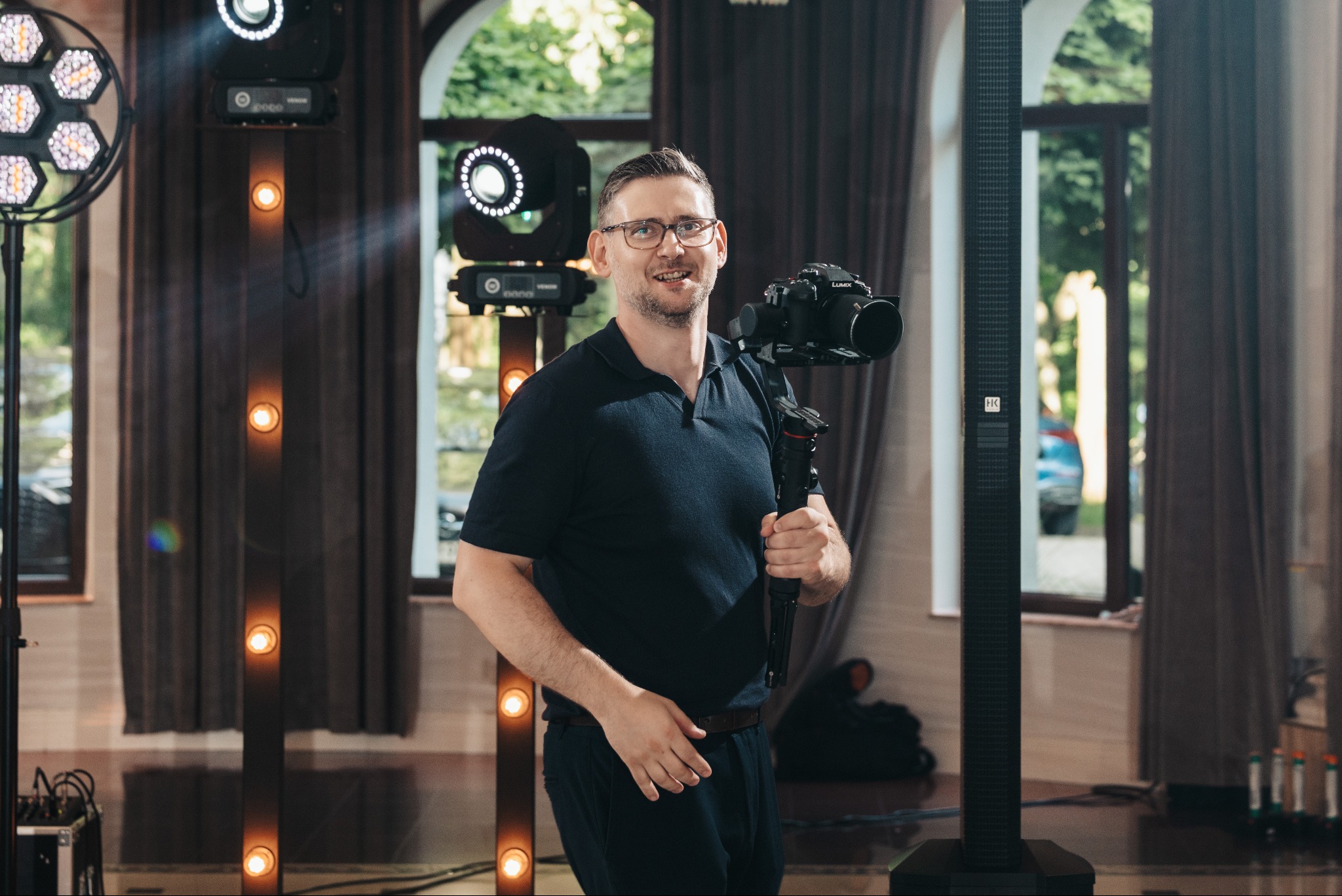 Uśmiechnięty mężczyzna w okularach trzyma kamerę Lumix na stabilizatorze, otoczony oświetleniem eventowym i kolumną nagłośnieniową HK Audio w eleganckim wnętrzu.