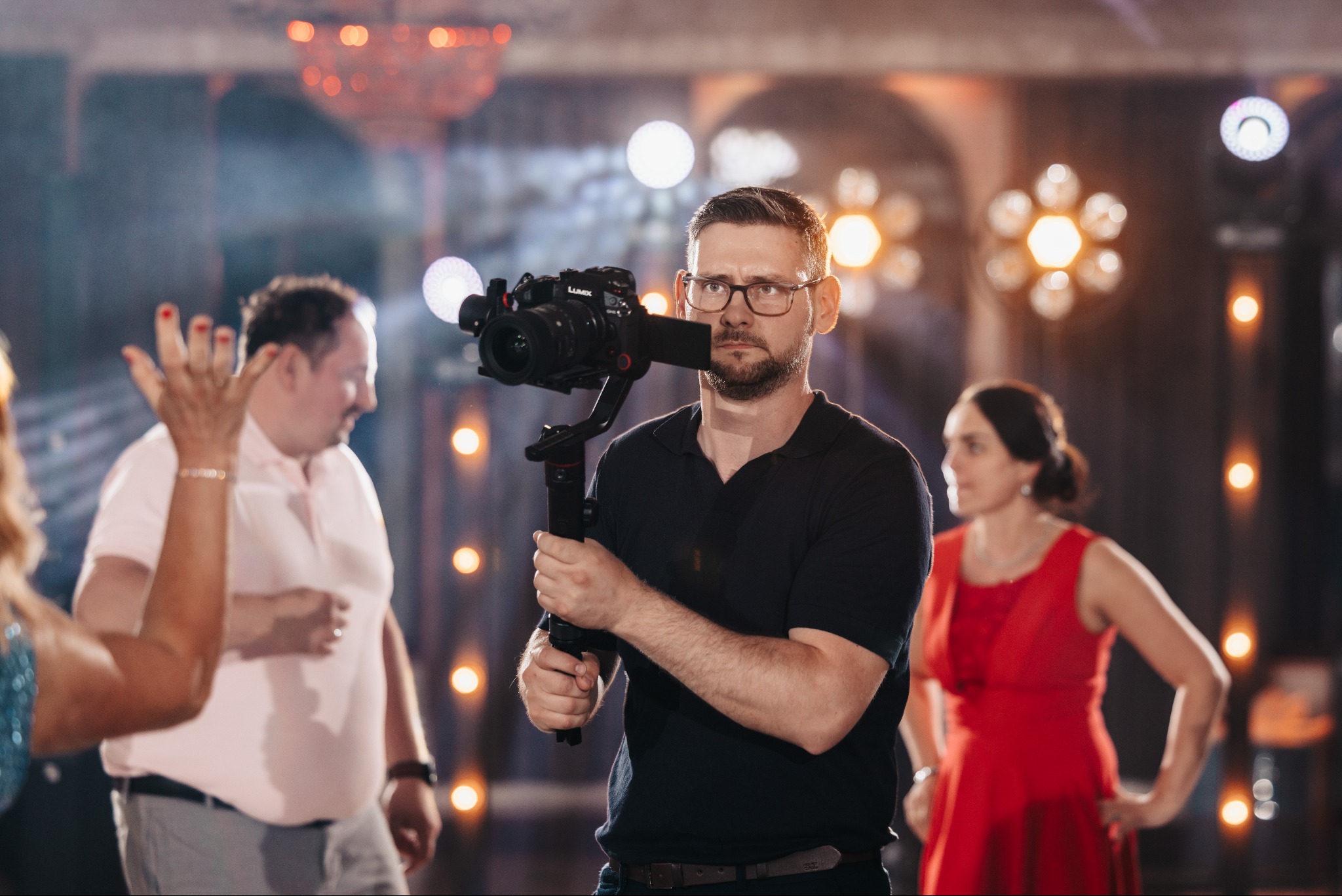 Kamerzysta z kamerą Lumix i stabilizatorem na tle oświetlonej sali, filmujący wydarzenie z gośćmi w tle. Drobne detale oświetlenia i stroju dodają unikalności.