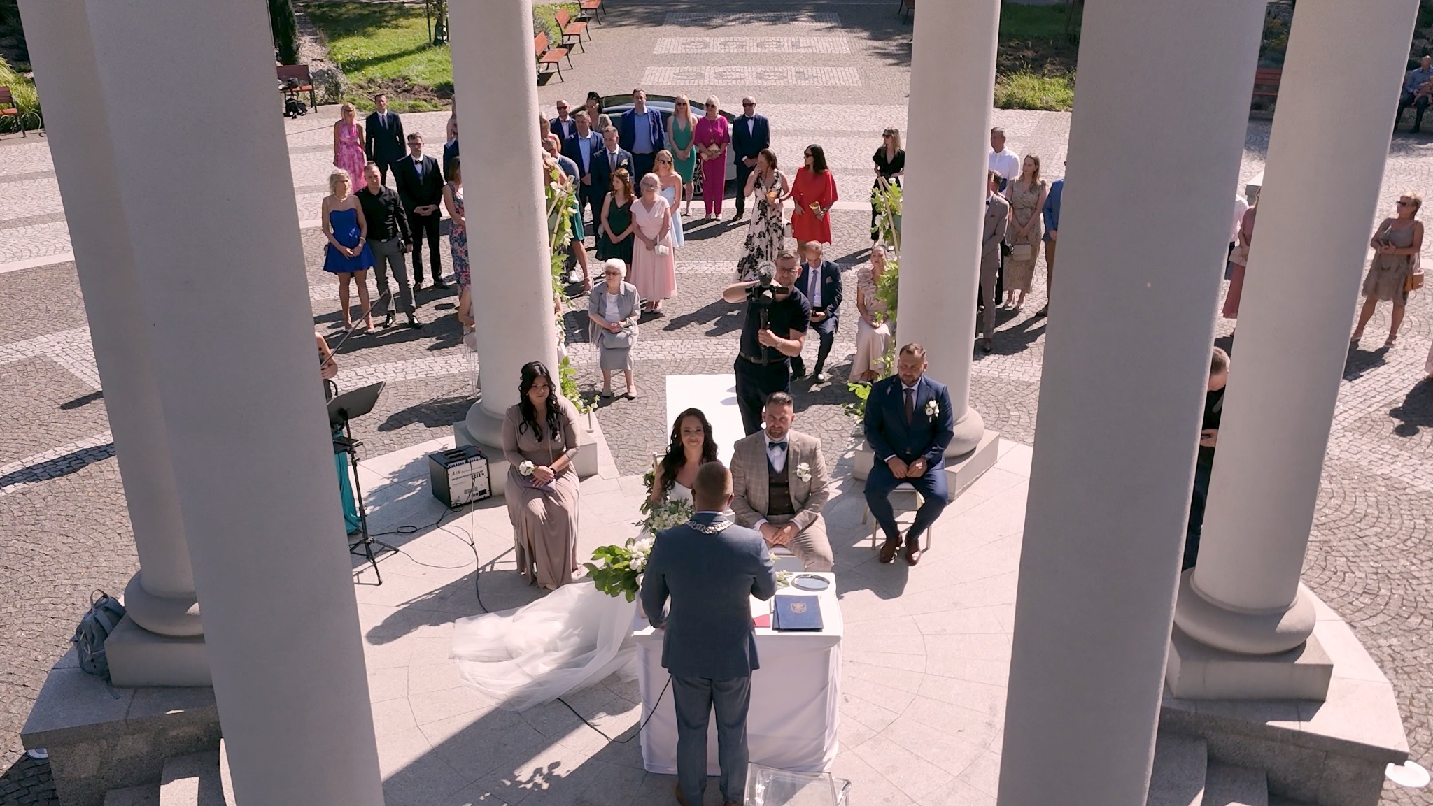 Uroczystość ślubna w plenerze, widok z góry przez kolumnadę, goście, para młoda, kamerzysta filmuje,  dekoracje kwiatowe.