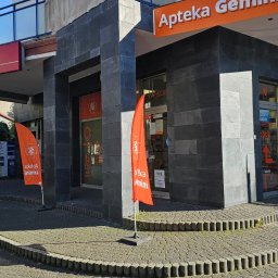 Kamień i Wnętrza - Elewacja apteki Gemini wykończona ciemnoszarą płytką imitującą kamień, z pomarańczowymi flagami reklamowymi i brukowanym chodnikiem.