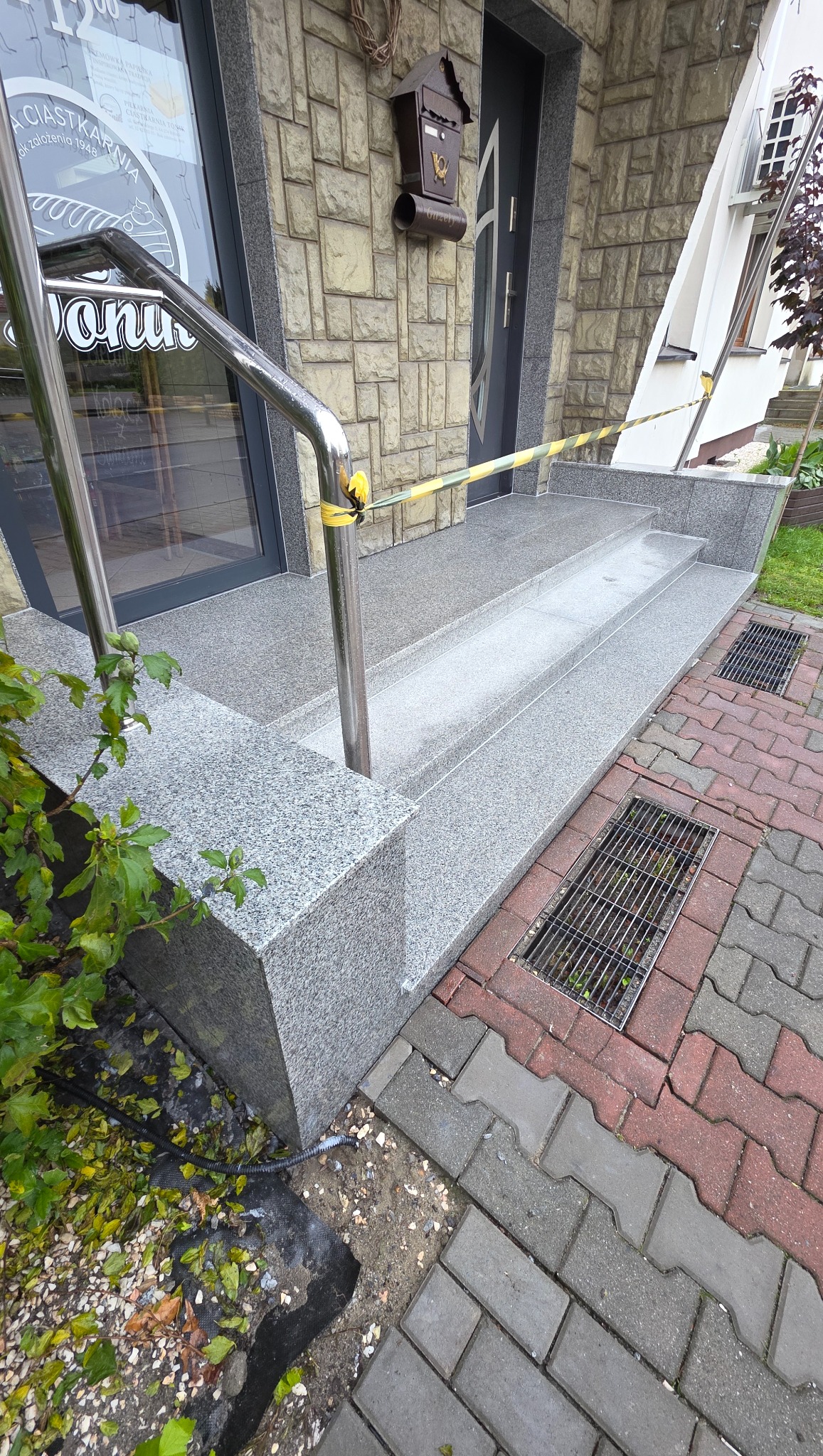 Granitowe schody wejściowe z metalową barierką, zabezpieczone taśmą ostrzegawczą. Widoczne detale wykończenia i kostka brukowa. Skrzynka pocztowa na elewacji.