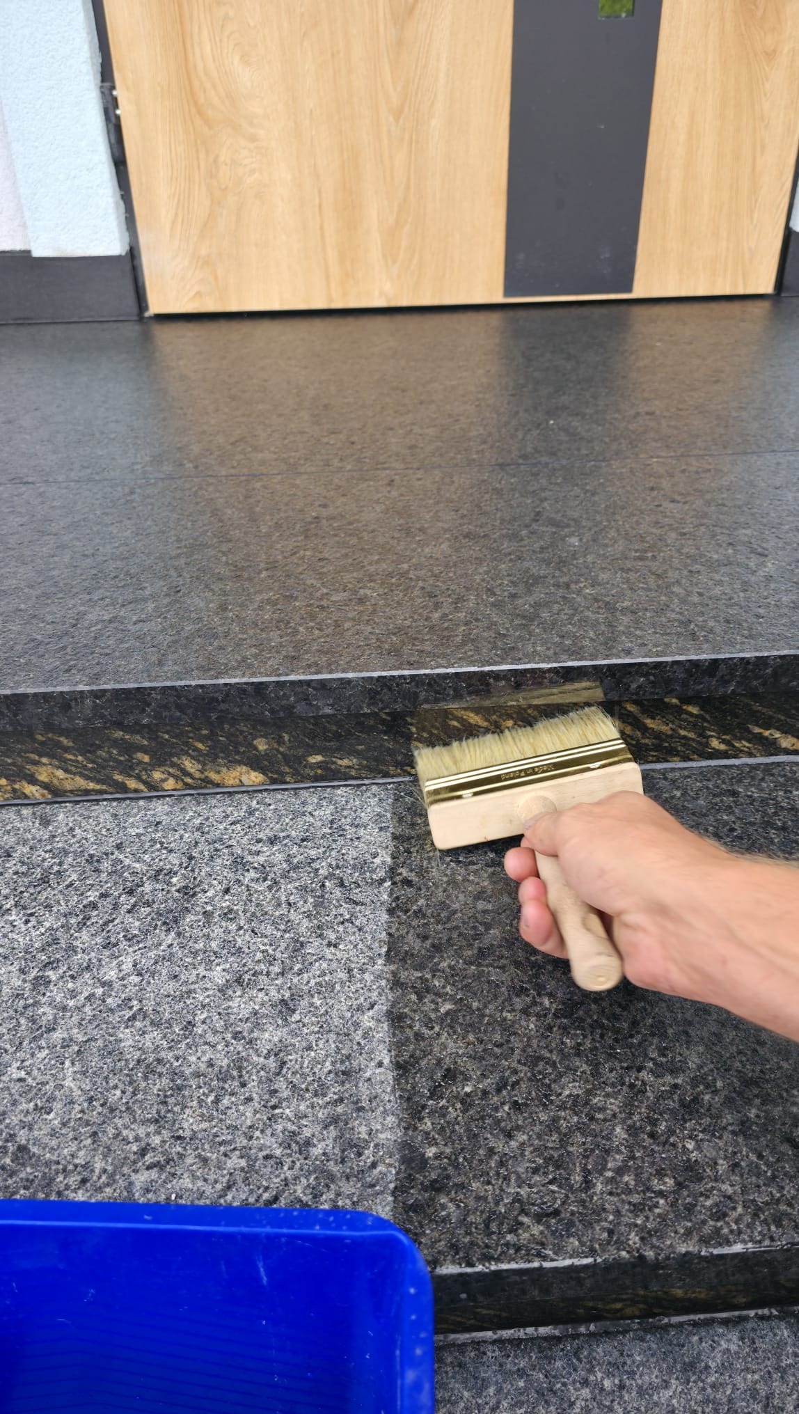 Granitowe stopnie po impregnacji. Widoczna różnica w kolorze po nałożeniu preparatu. Pędzel w dłoni aplikuje środek. W tle drzwi wejściowe i niebieskie wiadro.