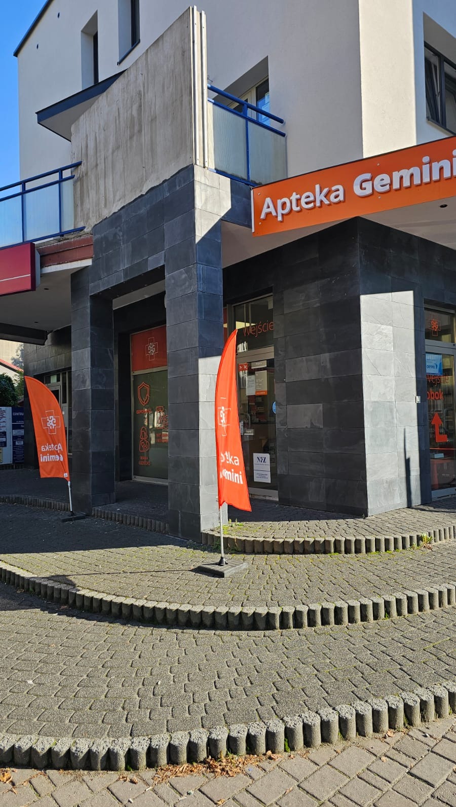 Elewacja apteki Gemini wykończona ciemnoszarą płytką imitującą kamień, z pomarańczowymi flagami reklamowymi i brukowanym chodnikiem.