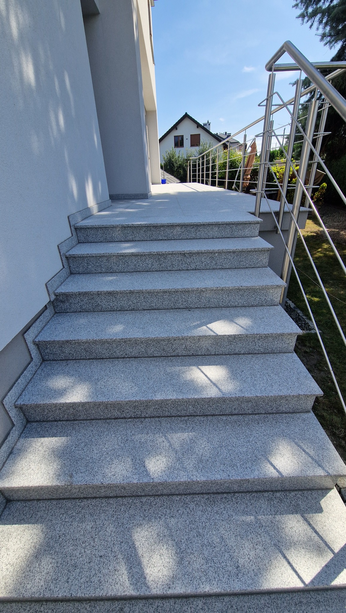Zewnętrzne schody z jasnoszarego kamienia z metalową balustradą, prowadzące do domu. Widok z dołu, w tle dom z dwuspadowym dachem i błękitne niebo.