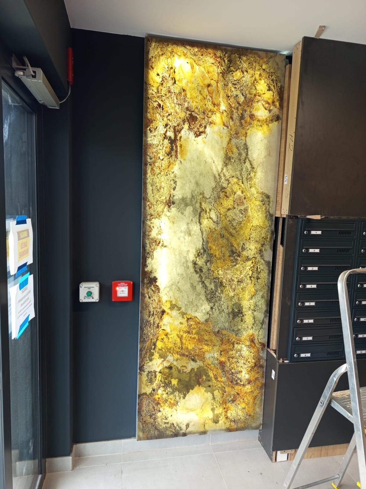 Dekoracyjna płyta ścienna z efektem złota i brązu, zamontowana w korytarzu obok skrzynek pocztowych, oświetlona od tyłu, tworząca unikalny element wykończenia wnętrza.