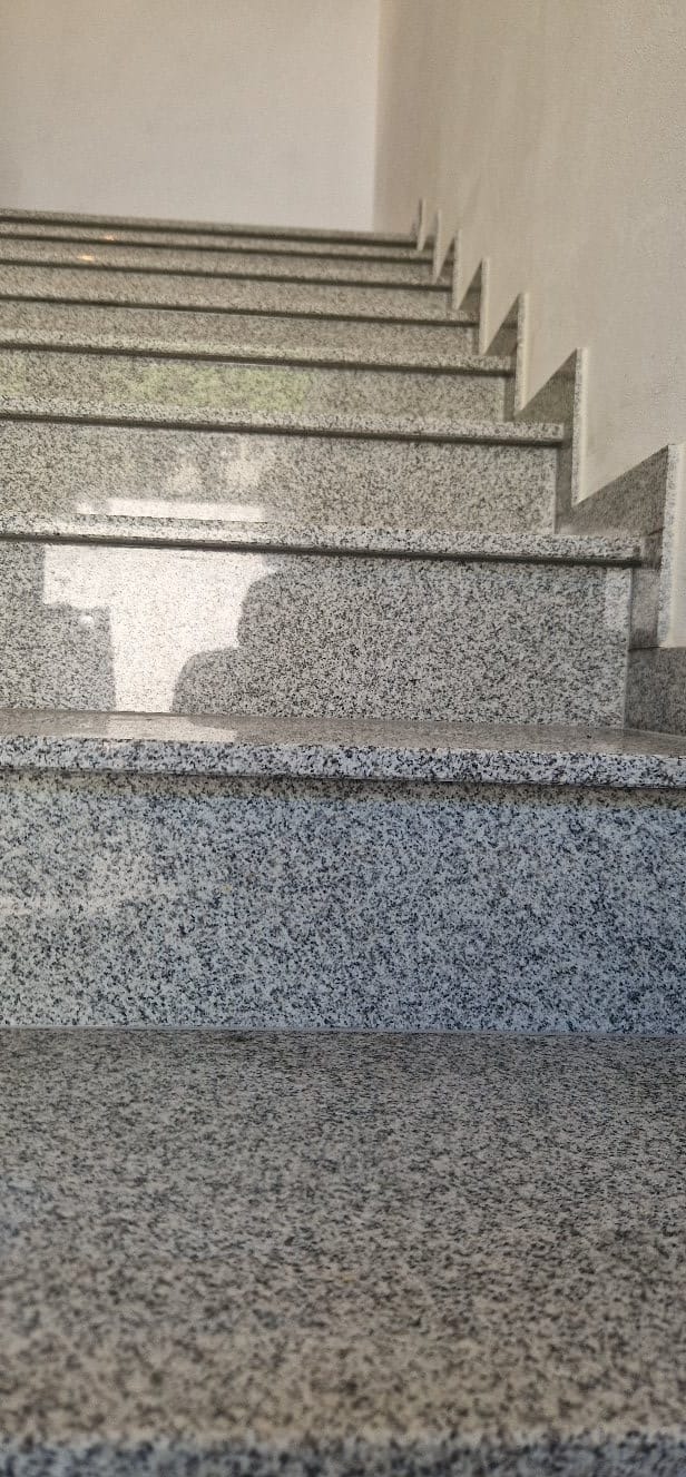Granitowe schody wewnętrzne z widoczną fakturą kamienia, oświetlone naturalnym światłem, z delikatnym cieniem osoby fotografującej na jednym ze stopni.