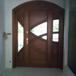 Drewniane drzwi wejściowe z łukowatym nadprożem i geometrycznymi przeszkleniami, złota klamka, widok z bliska na tle białych ścian.