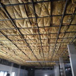 ROMAN CHERKASHYN USŁUGI OGÓLNOBUDOWLANE - Montaż izolacji z wełny mineralnej na metalowym ruszcie sufitowym w przestronnym, surowym wnętrzu budynku w trakcie remontu.