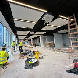 ROMAN CHERKASHYN USŁUGI OGÓLNOBUDOWLANE - Montaż sufitu podwieszanego w dużym pomieszczeniu biurowym z widocznymi pracownikami w odblaskowych kamizelkach, narzędziami i elementami konstrukcyjnymi na podłodze.