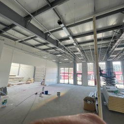ROMAN CHERKASHYN USŁUGI OGÓLNOBUDOWLANE - Wnętrze hali w trakcie prac wykończeniowych, widoczne elementy konstrukcji sufitu, materiały budowlane i podnośnik koszowy przy oknach.