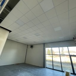 ROMAN CHERKASHYN USŁUGI OGÓLNOBUDOWLANE - Montaż sufitu kasetonowego w pomieszczeniu biurowym z dużymi oknami, widoczne niedokończone fragmenty i elementy konstrukcyjne.