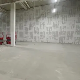 Przygotowania do montażu w dużej hali: ściana z widocznymi śladami po szpachlowaniu, podnośniki koszowe przy kolumnie nośnej, surowy beton na podłodze.