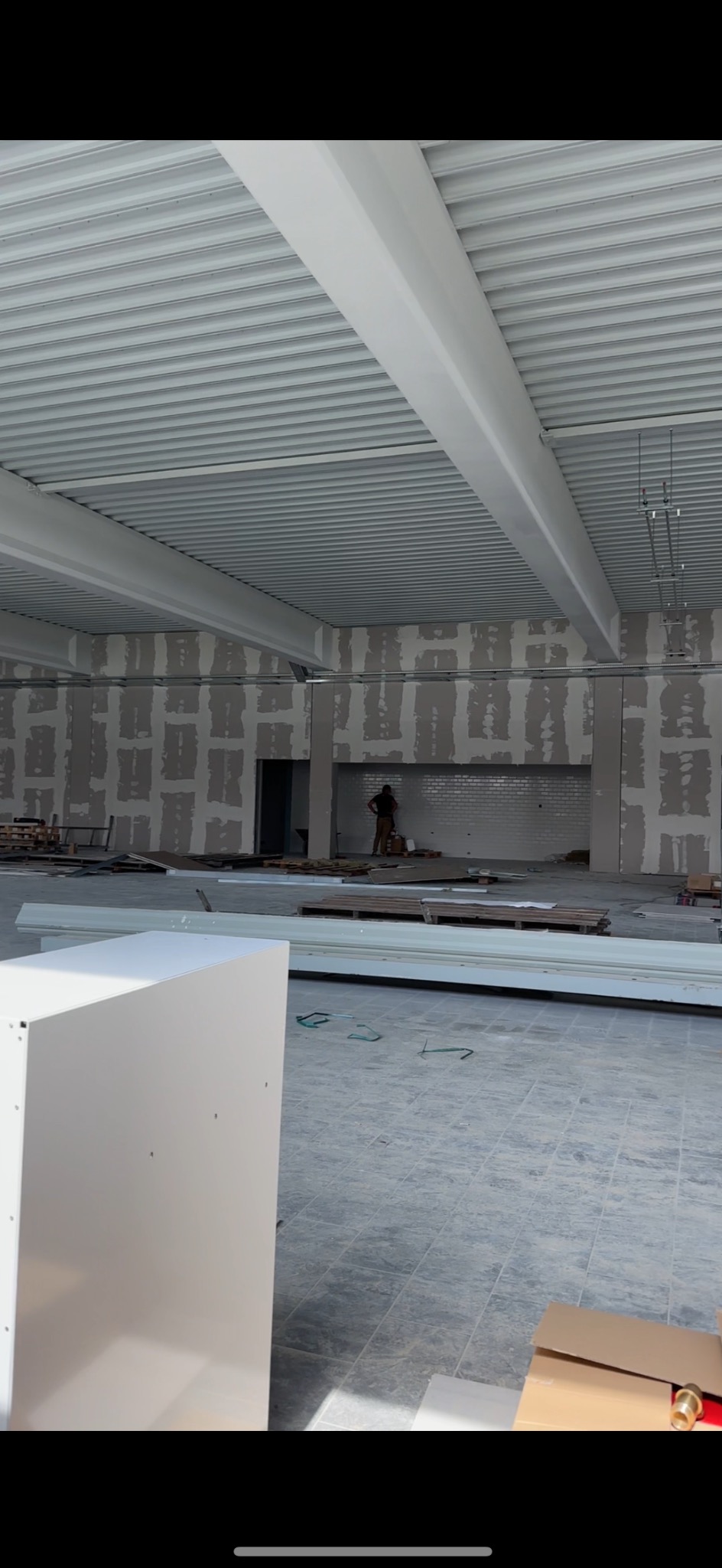 Wnętrze w trakcie remontu z widocznymi płytami na ścianach, metalowym sufitem podwieszanym i pracownikiem w tle.