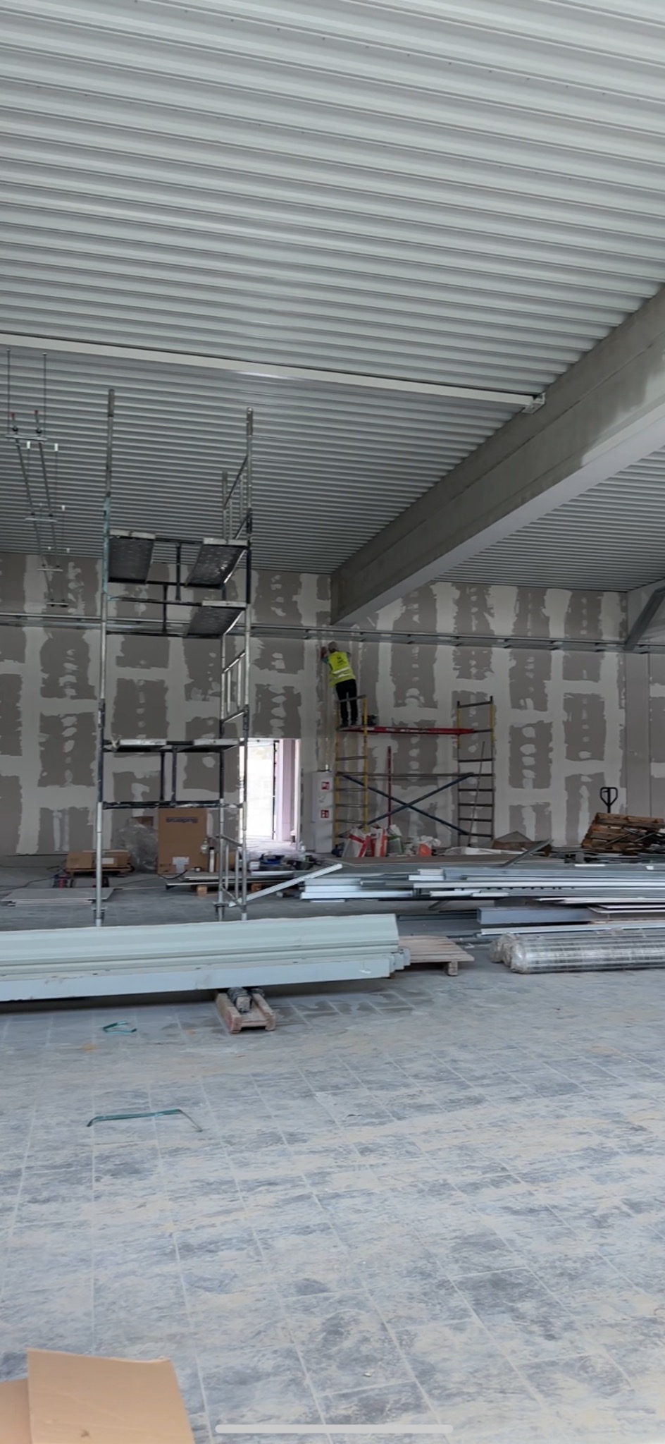 Montaż konstrukcji pod zabudowę w dużej hali, widoczne profile na ścianach, pracownik na rusztowaniu w kamizelce odblaskowej, sufit z blachy trapezowej.
