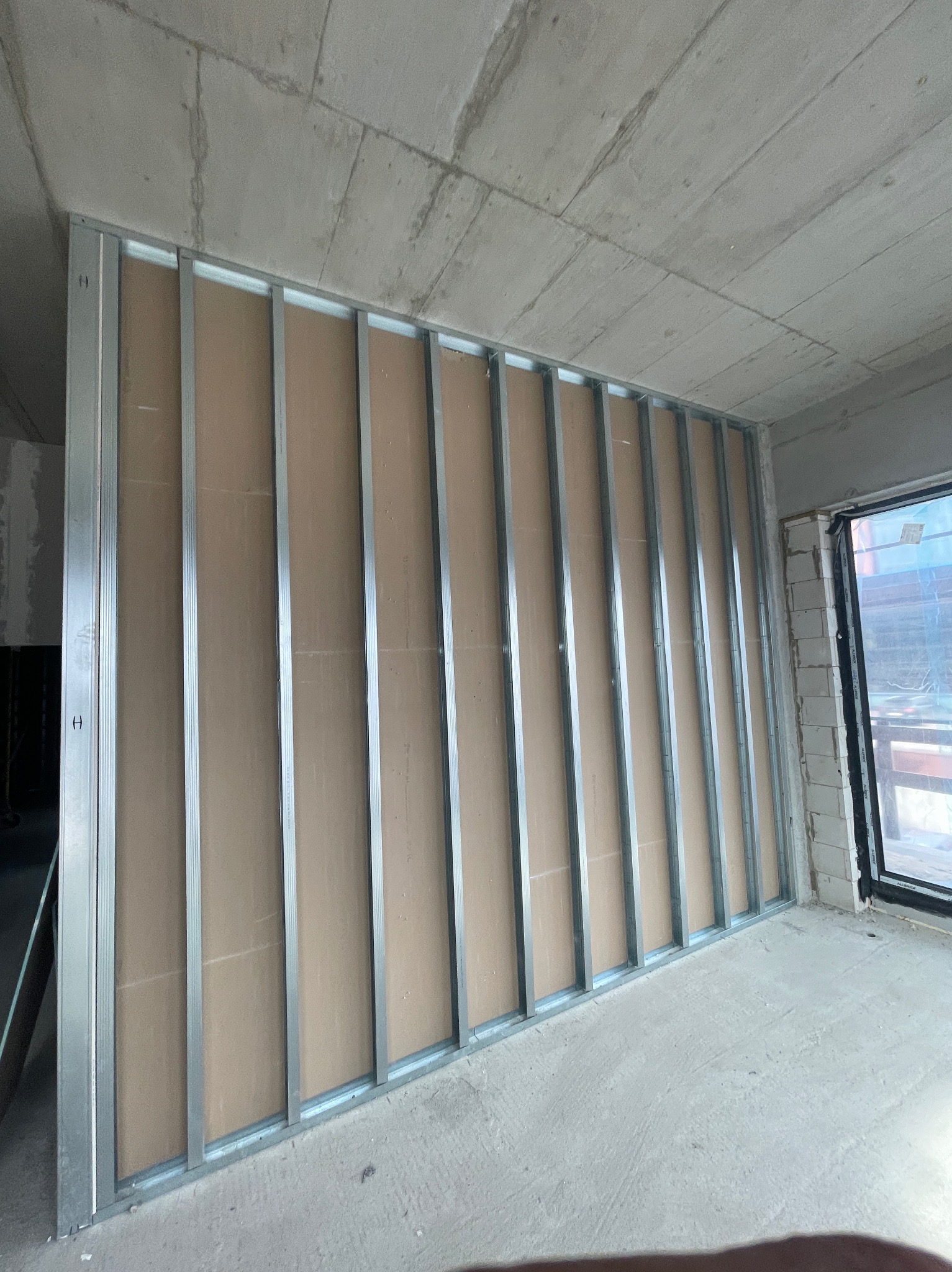 Stelaż z profili stalowych pod okładzinę w surowym wnętrzu, widoczne płyty w kolorze beżowym, surowy betonowy sufit i fragment okna.