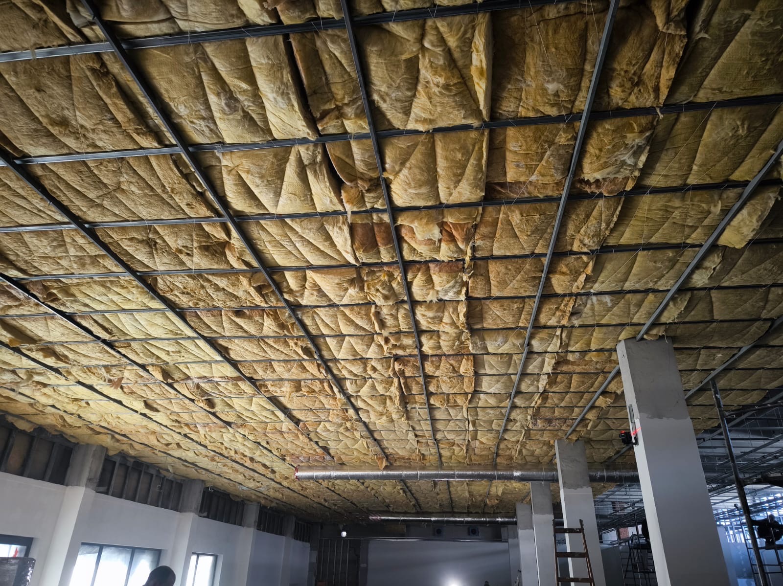 Montaż izolacji z wełny mineralnej na metalowym ruszcie sufitowym w przestronnym, surowym wnętrzu budynku w trakcie remontu.