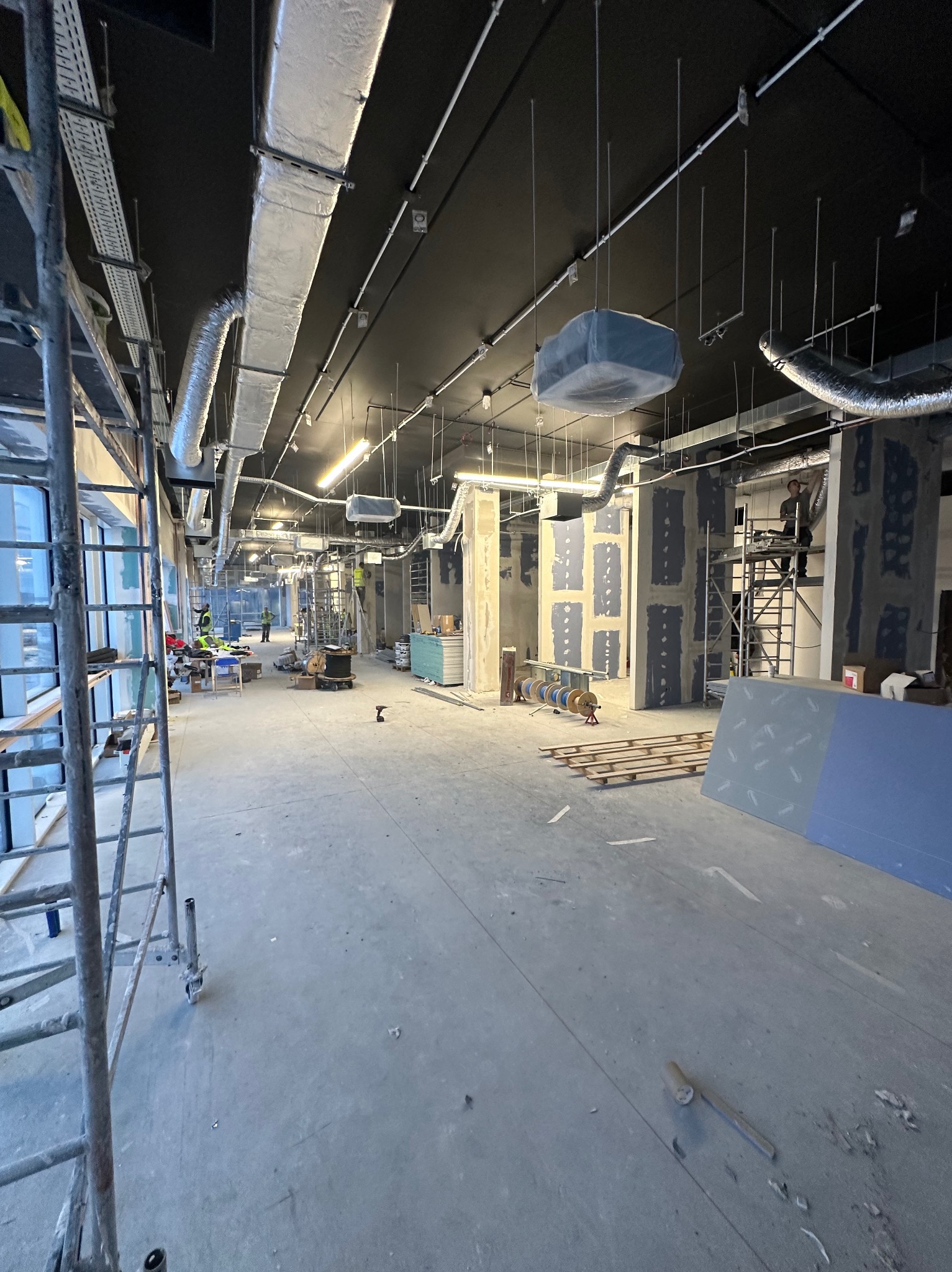 Wnętrze w trakcie remontu: widoczne szkielety ścian obłożone płytami, instalacje wentylacyjne pod czarnym sufitem, rusztowania i narzędzia budowlane na betonowej podłodze.