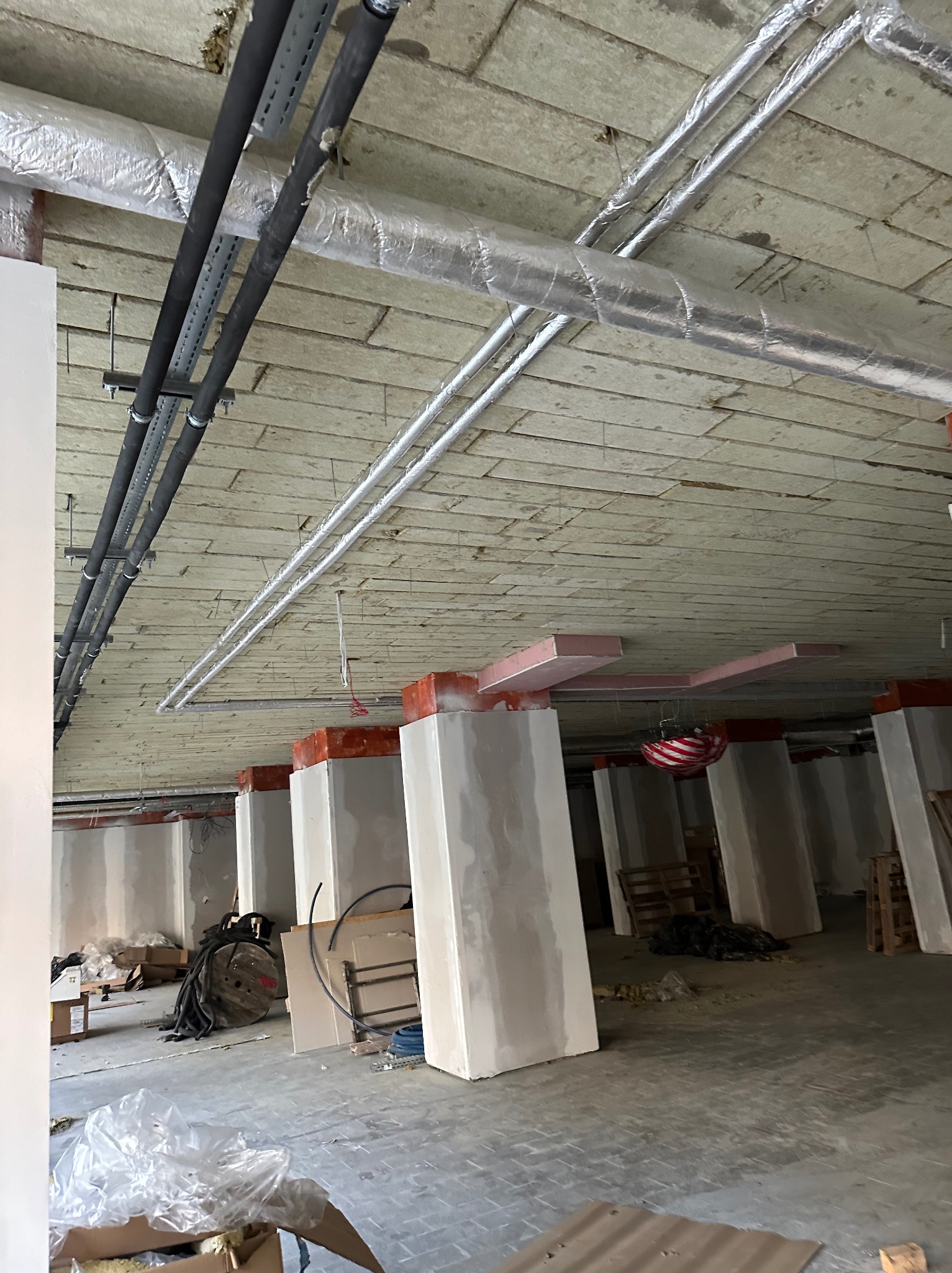 Surowy strop z izolacją, instalacjami i częściowo wykończonymi filarami w nowym budynku, widoczne materiały budowlane i narzędzia.