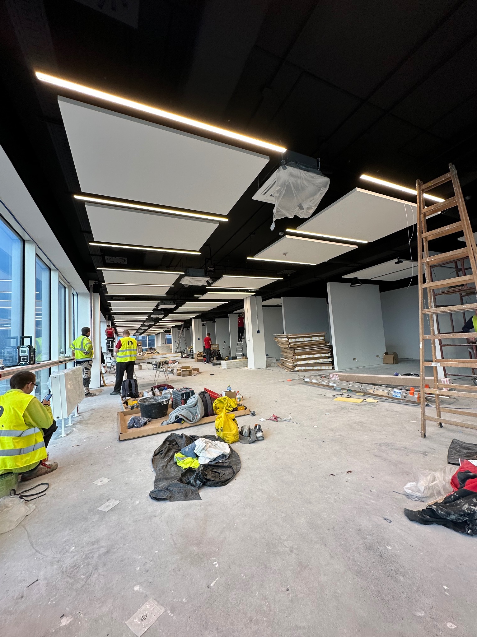Montaż sufitu podwieszanego w dużym pomieszczeniu biurowym z widocznymi pracownikami w odblaskowych kamizelkach, narzędziami i elementami konstrukcyjnymi na podłodze.