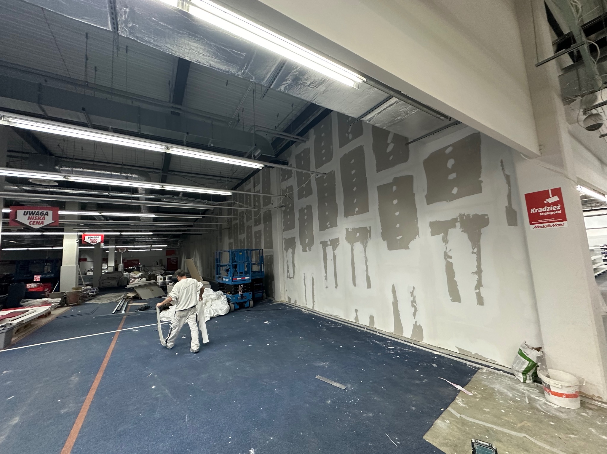 Montaż płyt na ścianie w sklepie: pracownik z profilami, surowa powierzchnia ścian z widocznymi łączeniami, podnośnik koszowy w tle, oświetlenie sklepowe.