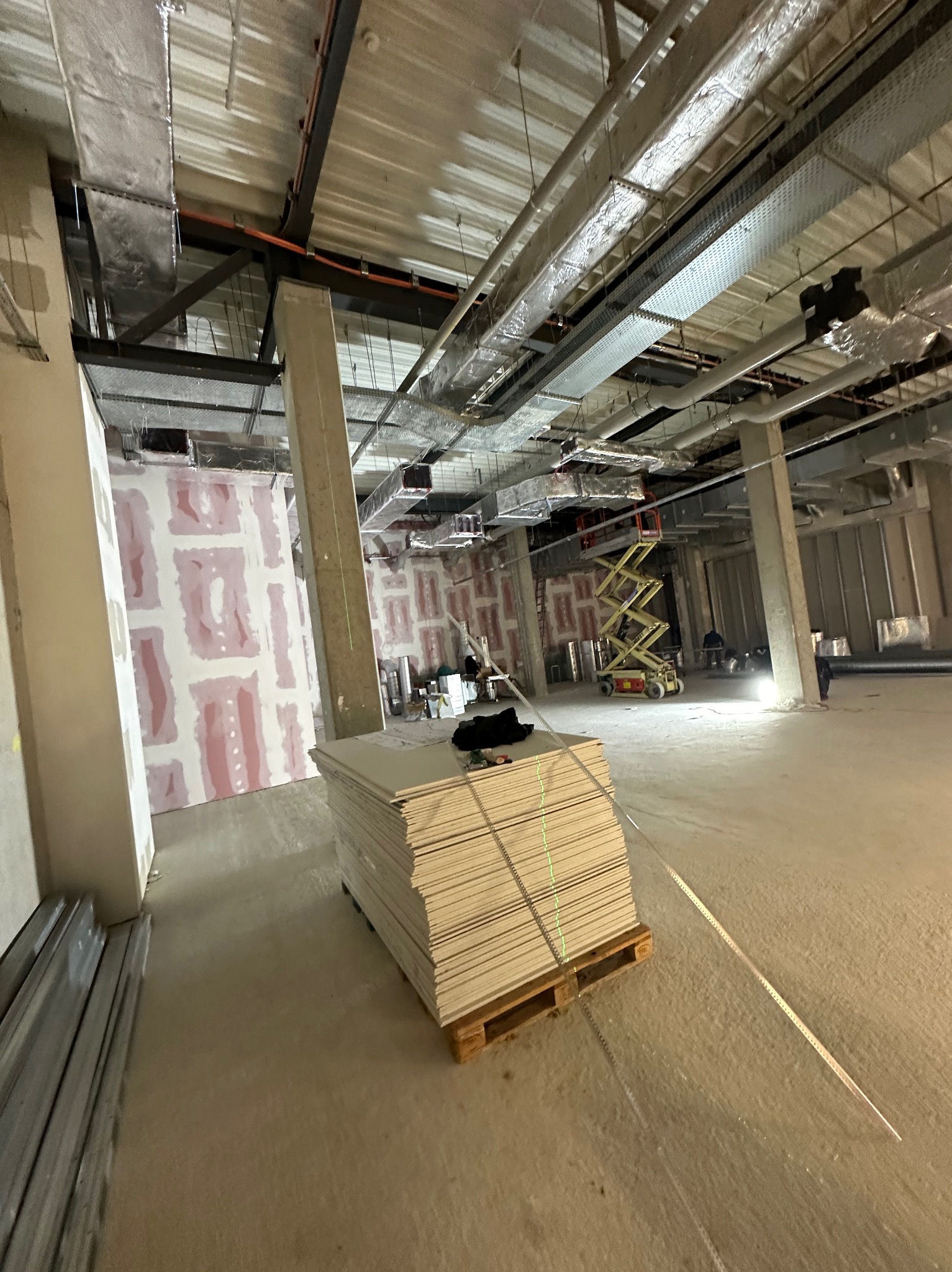 Stos płyt na palecie w surowym stanie budynku, widoczne profile metalowe i ściany z nałożoną warstwą gipsu, w tle podnośnik nożycowy.