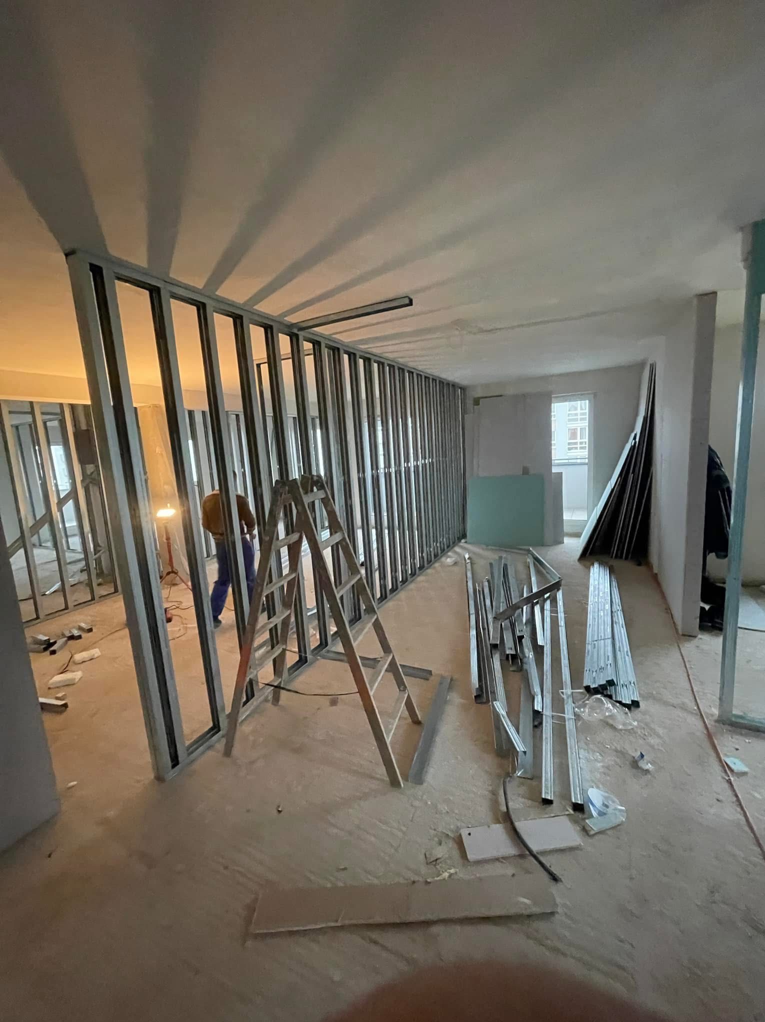 Wnętrze w trakcie montażu metalowej konstrukcji ścianki działowej, widoczna drabina, profile oraz pracownik w oddali przy lampie.