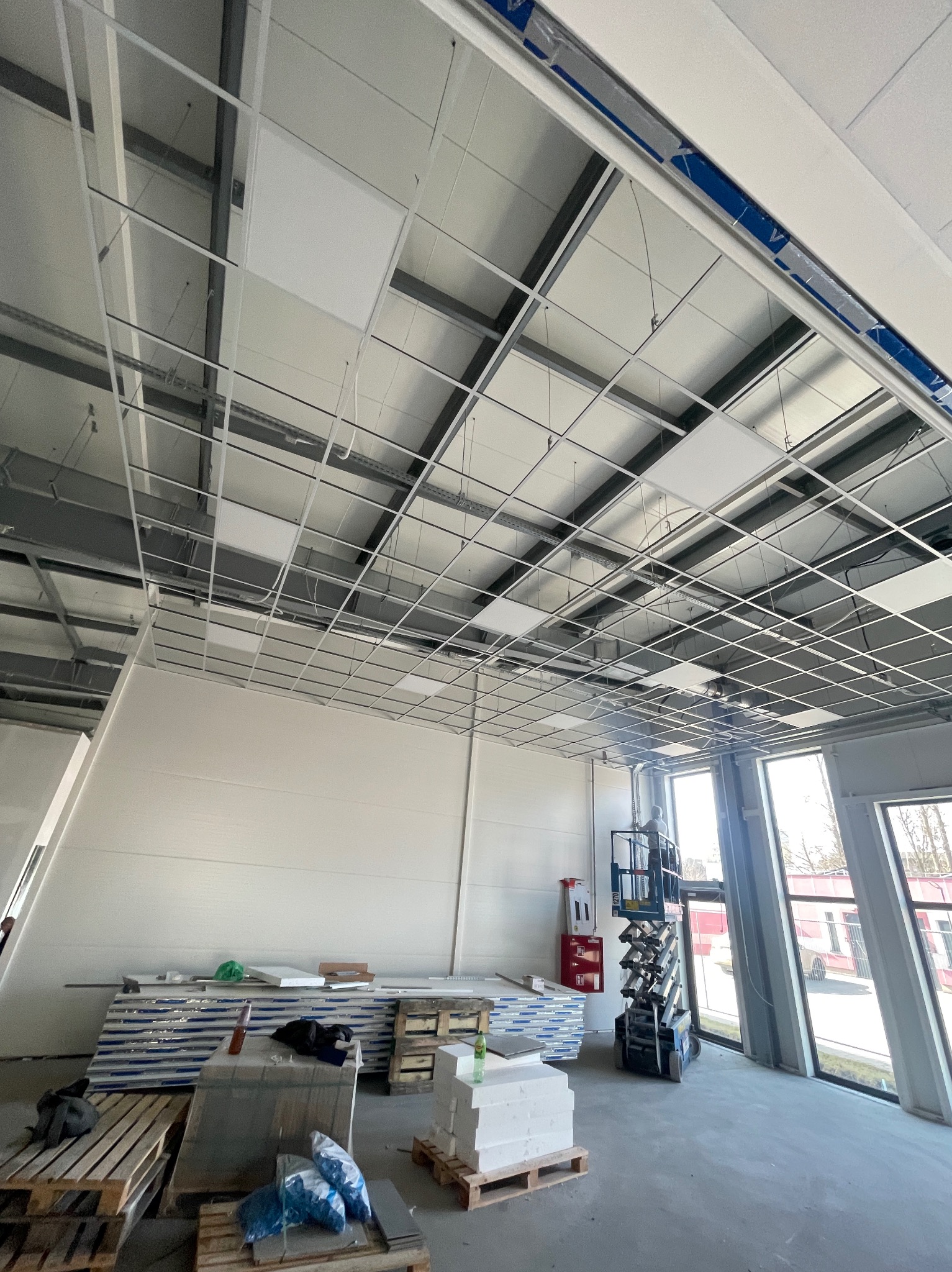 Montaż sufitu podwieszanego z płyt karton-gips w hali przemysłowej: widoczna metalowa konstrukcja, częściowo zamontowane płyty, podnośnik nożycowy i palety z materiałami.