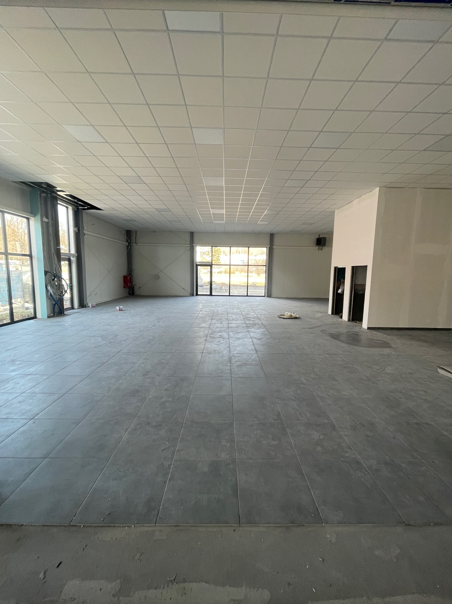 Wnętrze przestronnego pomieszczenia w trakcie wykańczania z widocznym sufitem kasetonowym, surowymi ścianami i szarą podłogą z płytek. Widoczne okna z metalowymi ramami oraz częściowo wykończony...