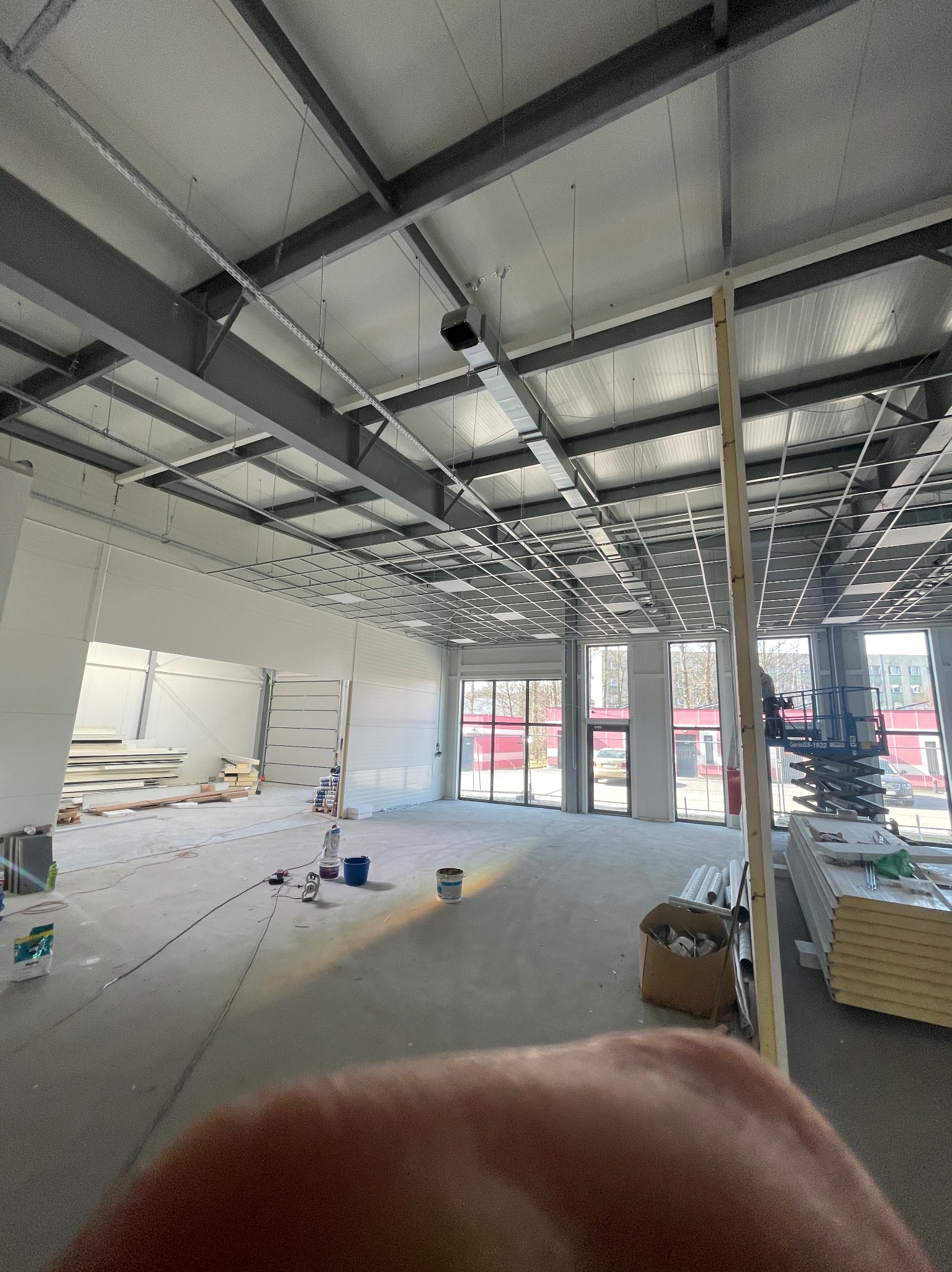 Wnętrze hali w trakcie prac wykończeniowych, widoczne elementy konstrukcji sufitu, materiały budowlane i podnośnik koszowy przy oknach.