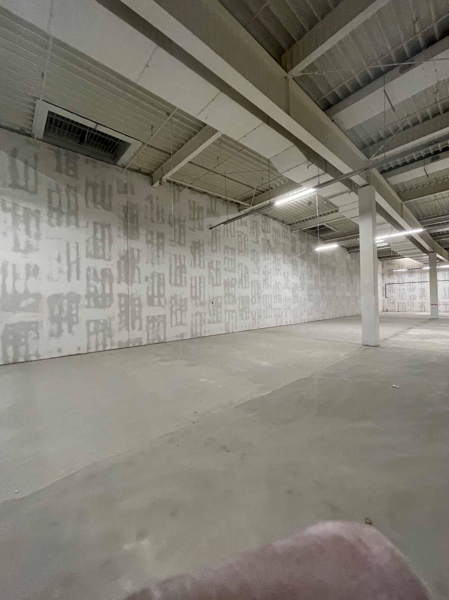Wnętrze hali przemysłowej w trakcie wykańczania ścian, widoczne ślady po szpachlowaniu na surowych ścianach, sufit z widoczną konstrukcją i oświetleniem.