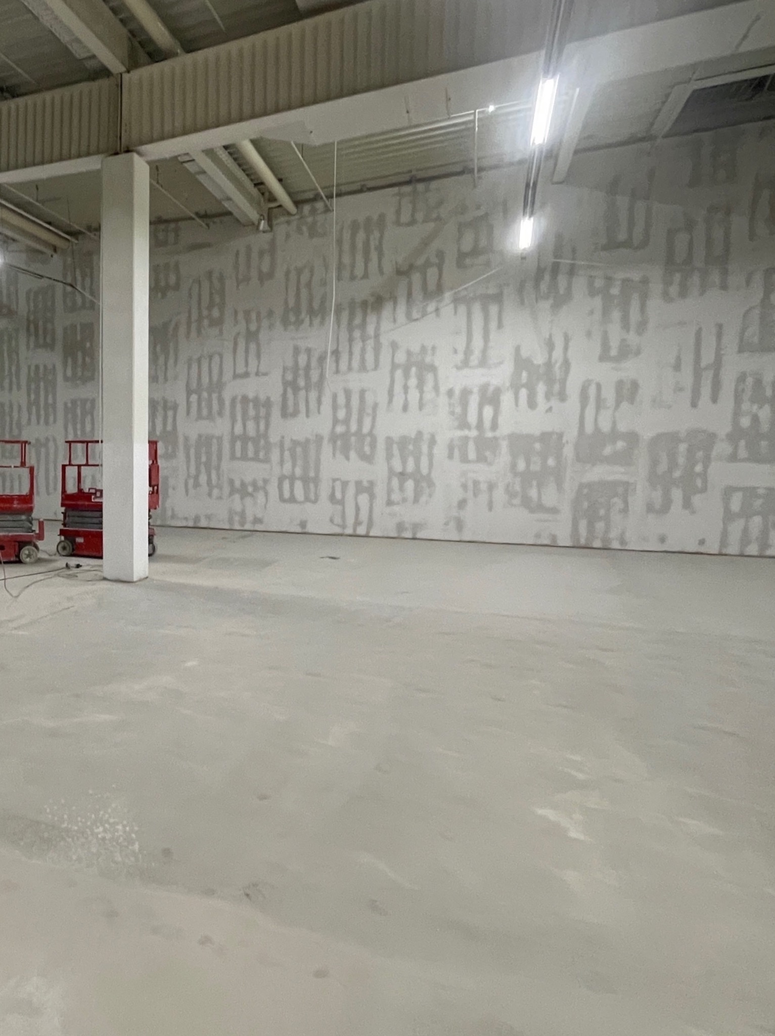 Przygotowania do montażu w dużej hali: ściana z widocznymi śladami po szpachlowaniu, podnośniki koszowe przy kolumnie nośnej, surowy beton na podłodze.