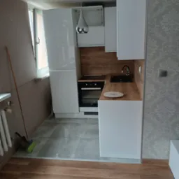 Nowoczesna kuchnia po remoncie z białymi szafkami, drewnianym blatem i czarnym zlewem; na podłodze szare płytki, widoczna miotła przy grzejniku, ściana z tapetą w kwiaty.