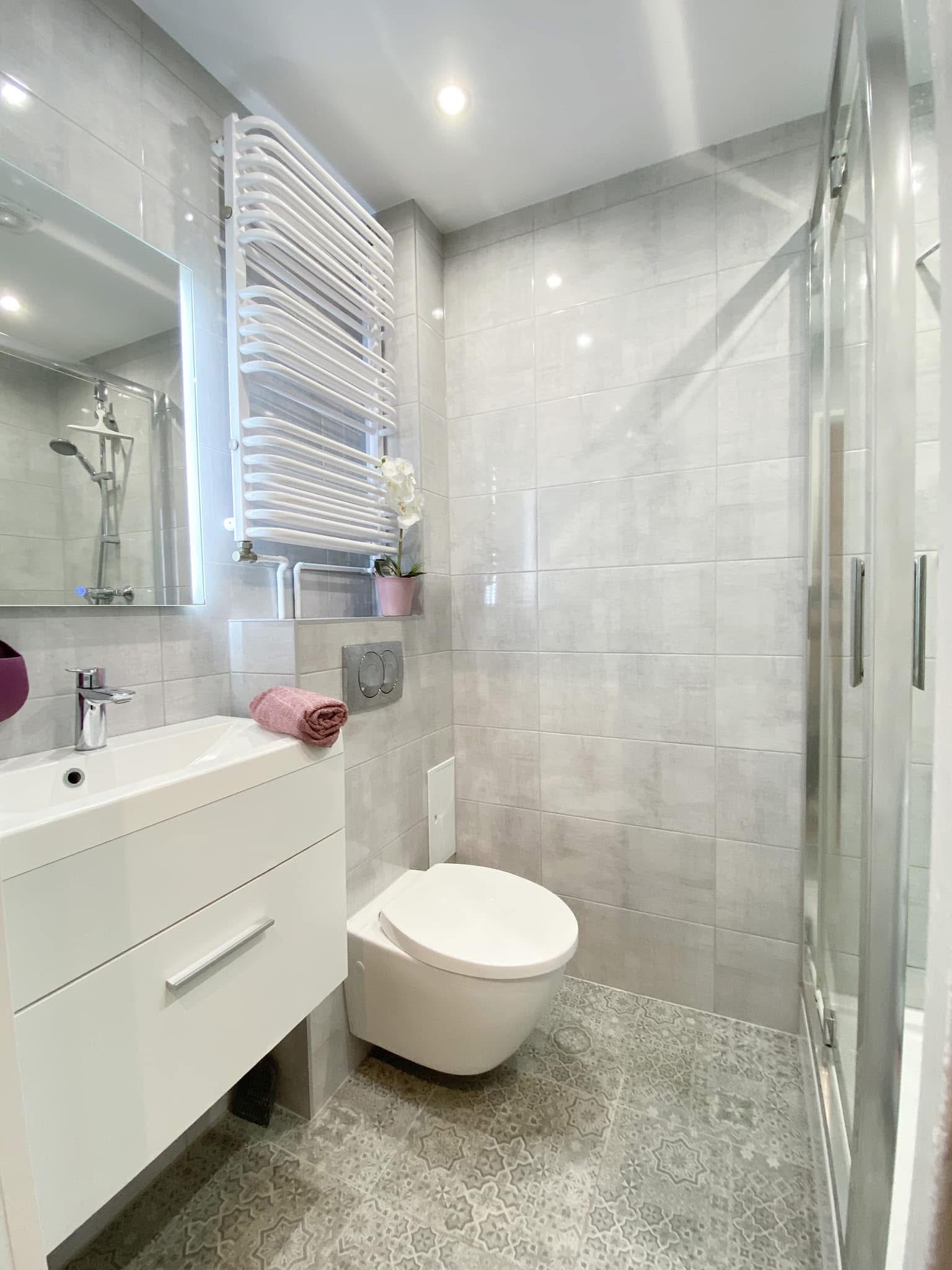 Jasnoszara łazienka po remoncie z kabiną prysznicową, umywalką z szafką, toaletą podwieszaną i grzejnikiem drabinkowym; na podłodze wzorzyste płytki.