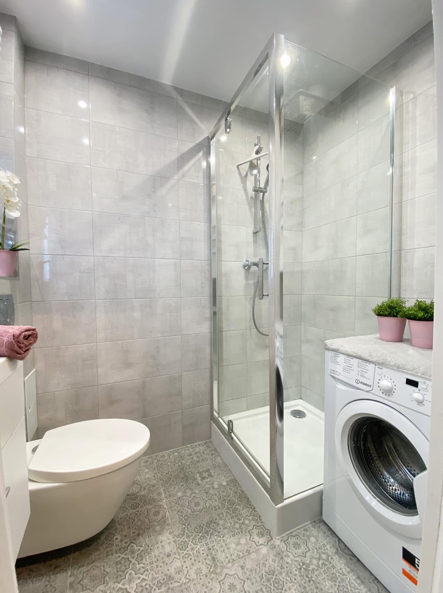 Jasna łazienka po remoncie z kabiną prysznicową, wiszącą toaletą, pralką Indesit i wzorzystymi szarymi płytkami podłogowymi.