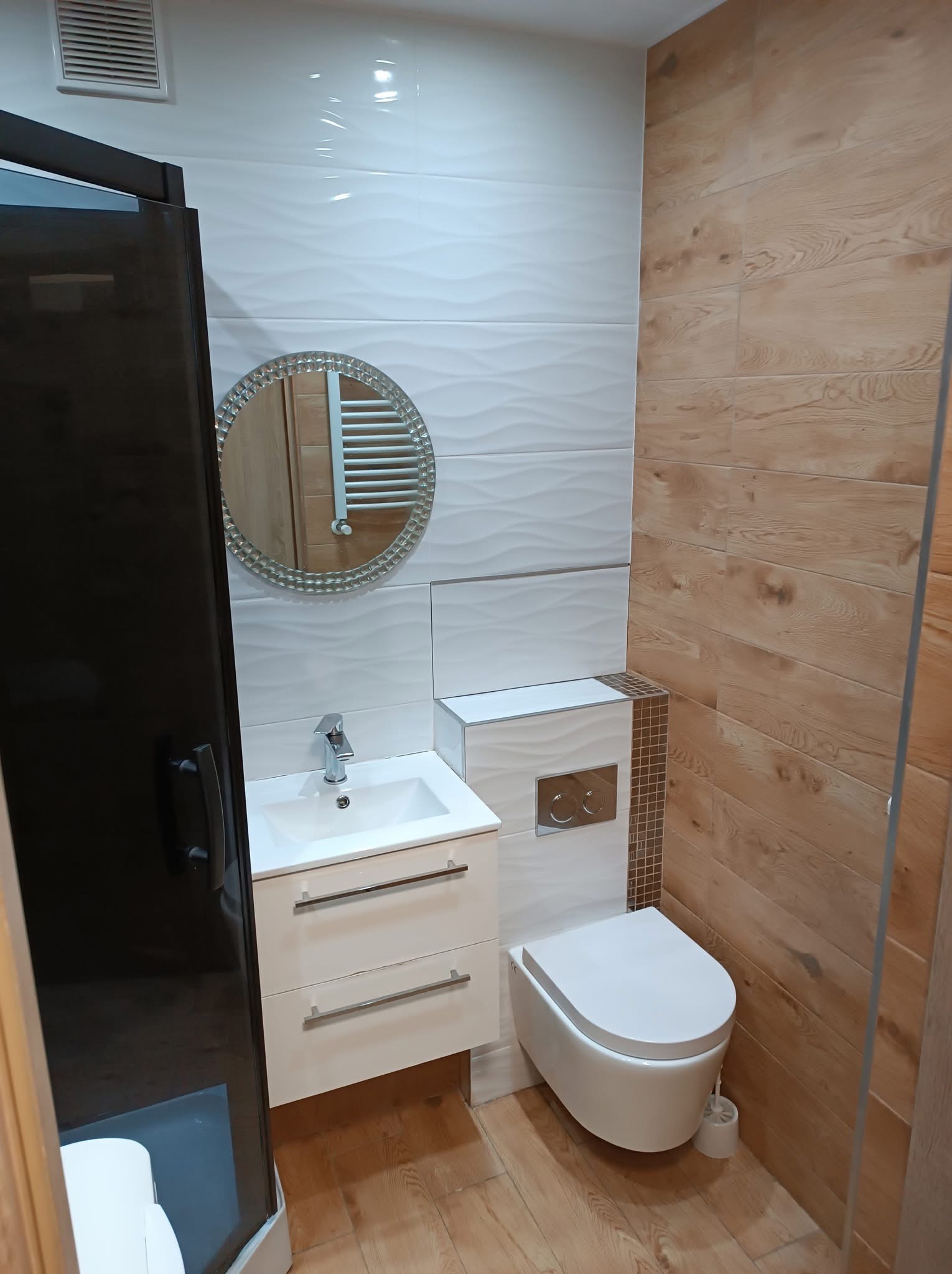 Nowoczesna łazienka z białymi płytkami o falistej strukturze, drewnopodobnymi panelami ściennymi, okrągłym lustrem z dekoracyjną ramą, podwieszaną toaletą i czarną kabiną prysznicową.