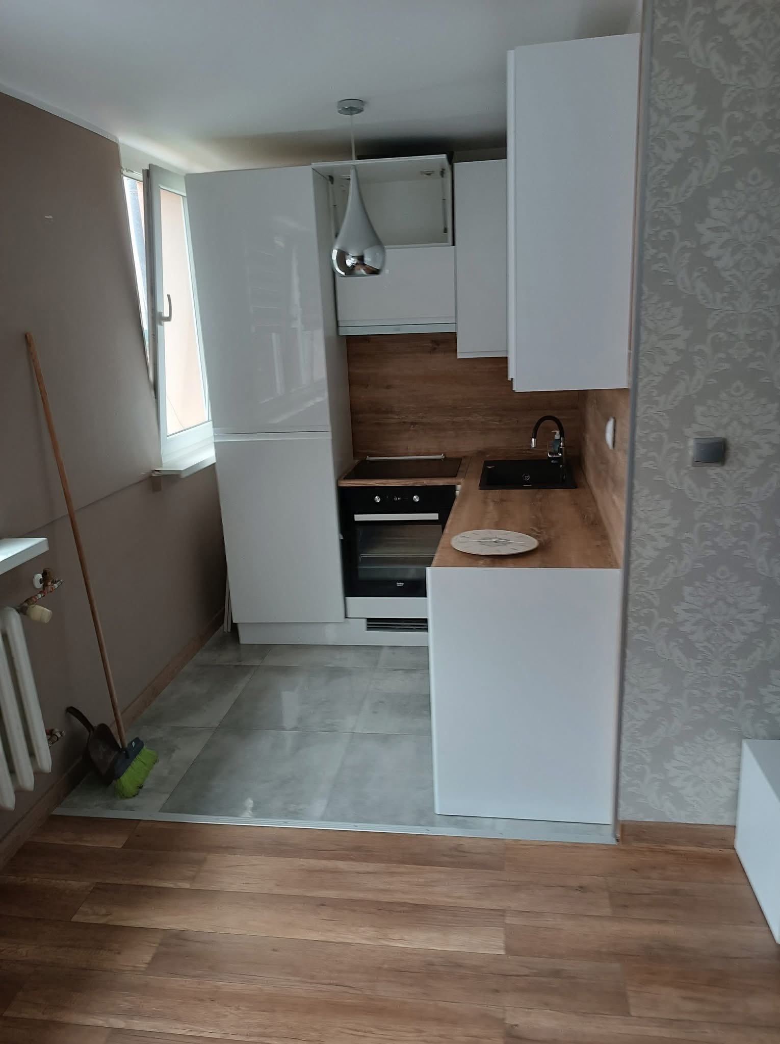 Nowoczesna kuchnia po remoncie z białymi szafkami, drewnianym blatem i czarnym zlewem; na podłodze szare płytki, widoczna miotła przy grzejniku, ściana z tapetą w kwiaty.