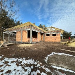 RAMSTROI Sp. z o.o. - Wznoszenie ścian z czerwonej cegły na parterowym domu jednorodzinnym z widocznymi elementami betonowymi, częściowo odeskowanym dachem i resztkami śniegu na ziemi.