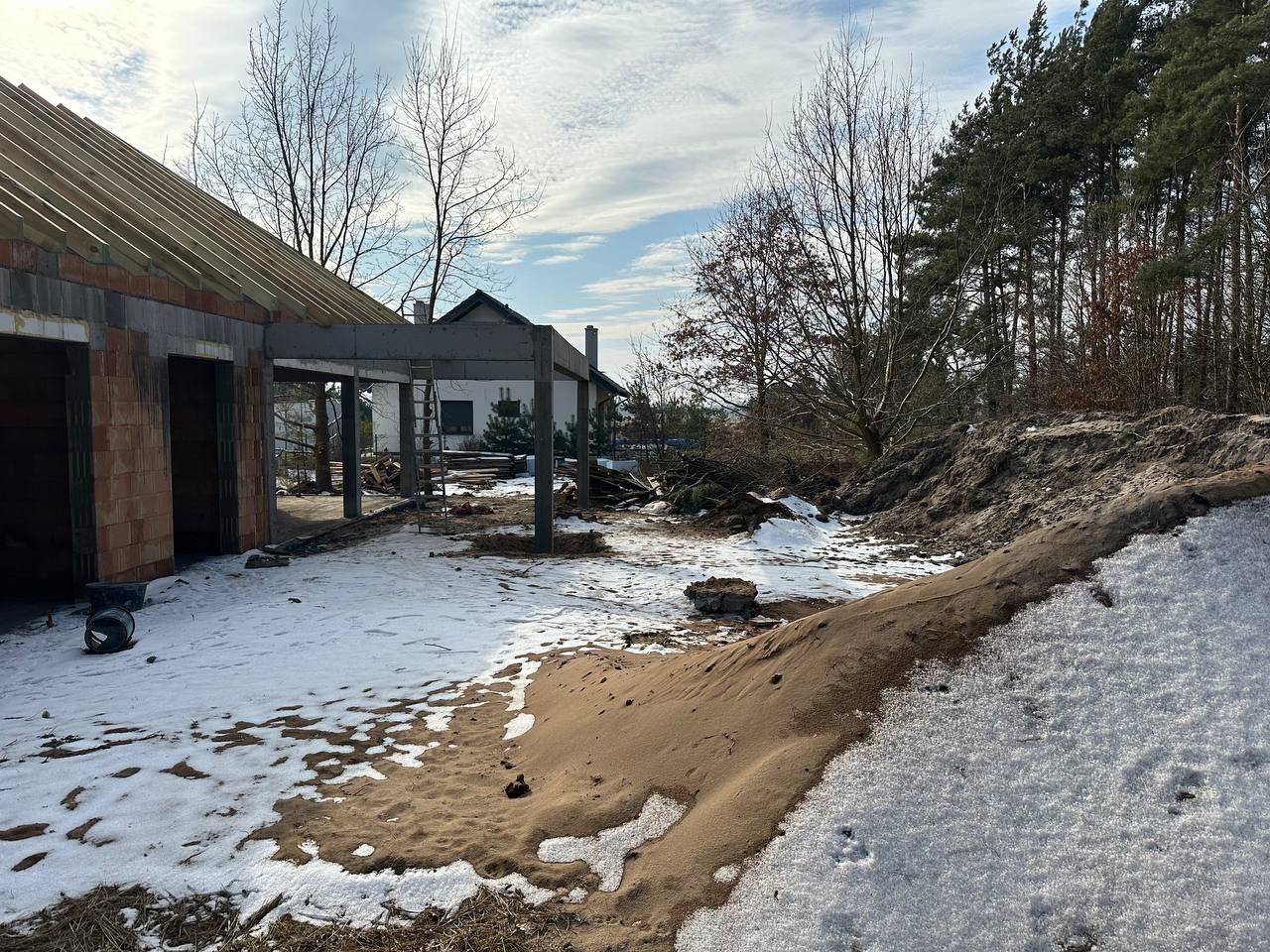 Wczesny etap budowy domu z czerwonej cegły i żelbetowymi elementami konstrukcyjnymi w zimowym krajobrazie; widoczny szkielet budynku, częściowo ośnieżony teren i las w tle.