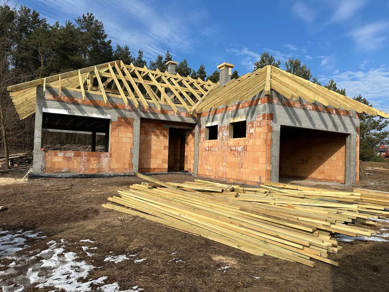 Budowa domu jednorodzinnego z czerwonej cegły, widoczny szkielet dachu z drewnianych belek, na pierwszym planie stos drewna budowlanego, w tle las i błękitne niebo.