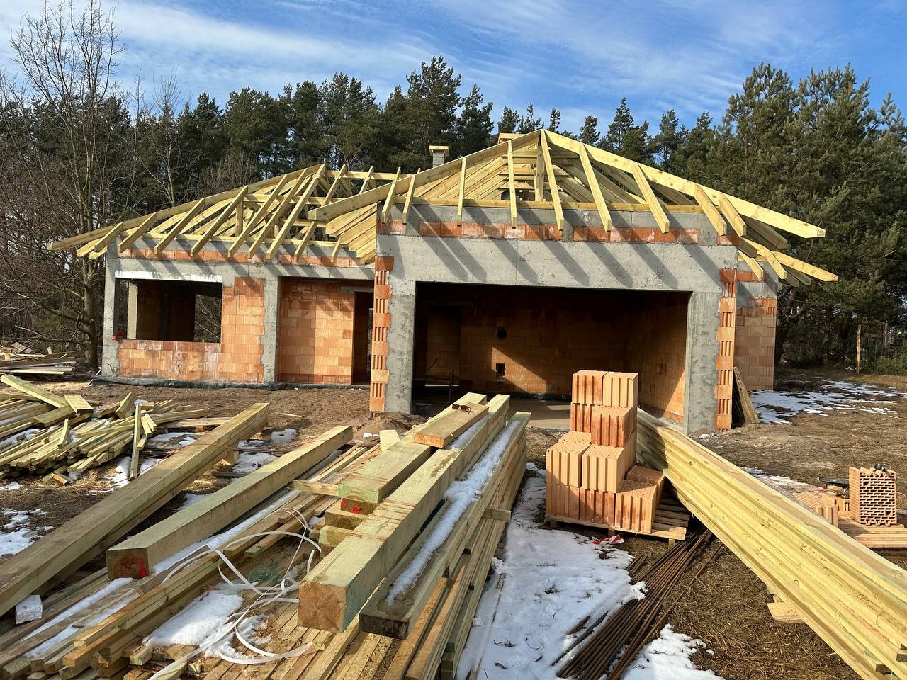 Surowy stan domu z cegły z drewnianą konstrukcją dachu w trakcie budowy, widoczne materiały budowlane i resztki śniegu na ziemi.