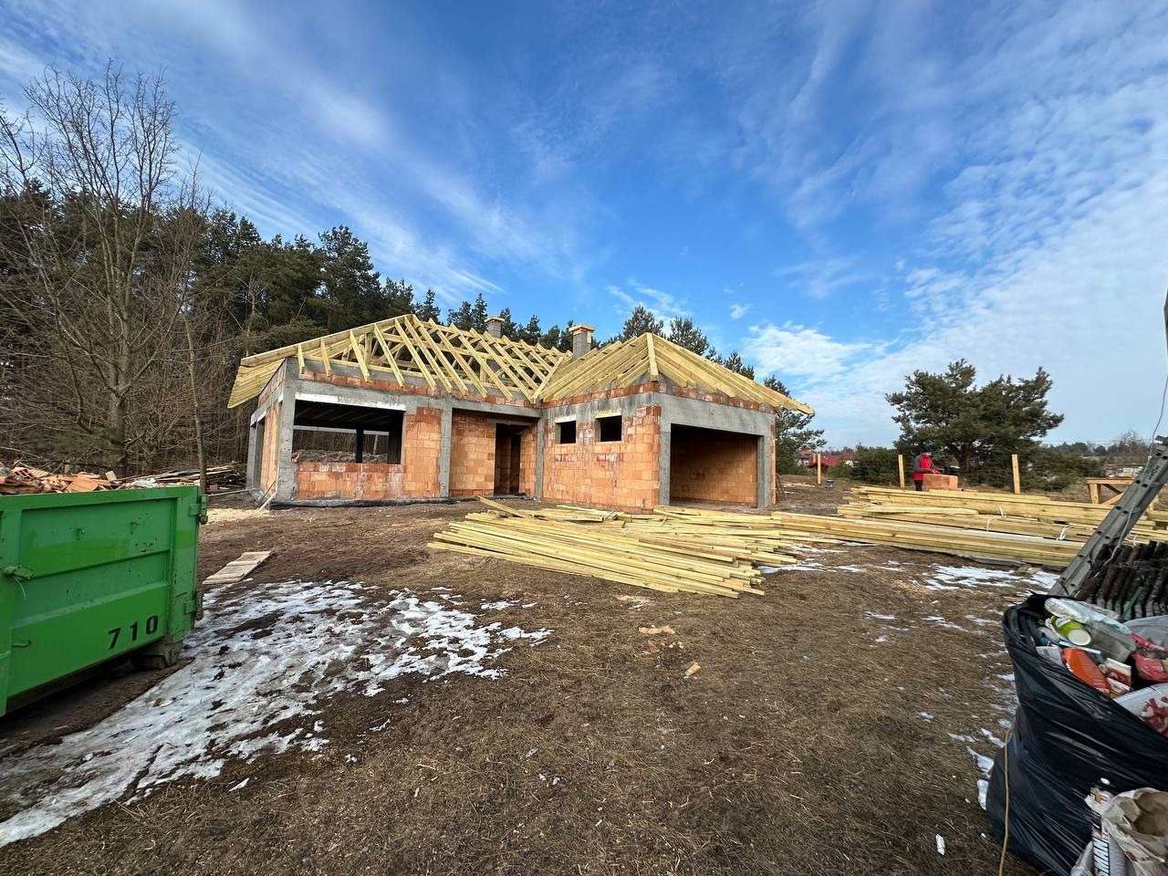 Budowa domu jednorodzinnego z czerwonej cegły w trakcie realizacji, widoczne drewniane elementy konstrukcji dachu, zielony kontener na odpady i resztki śniegu na ziemi.