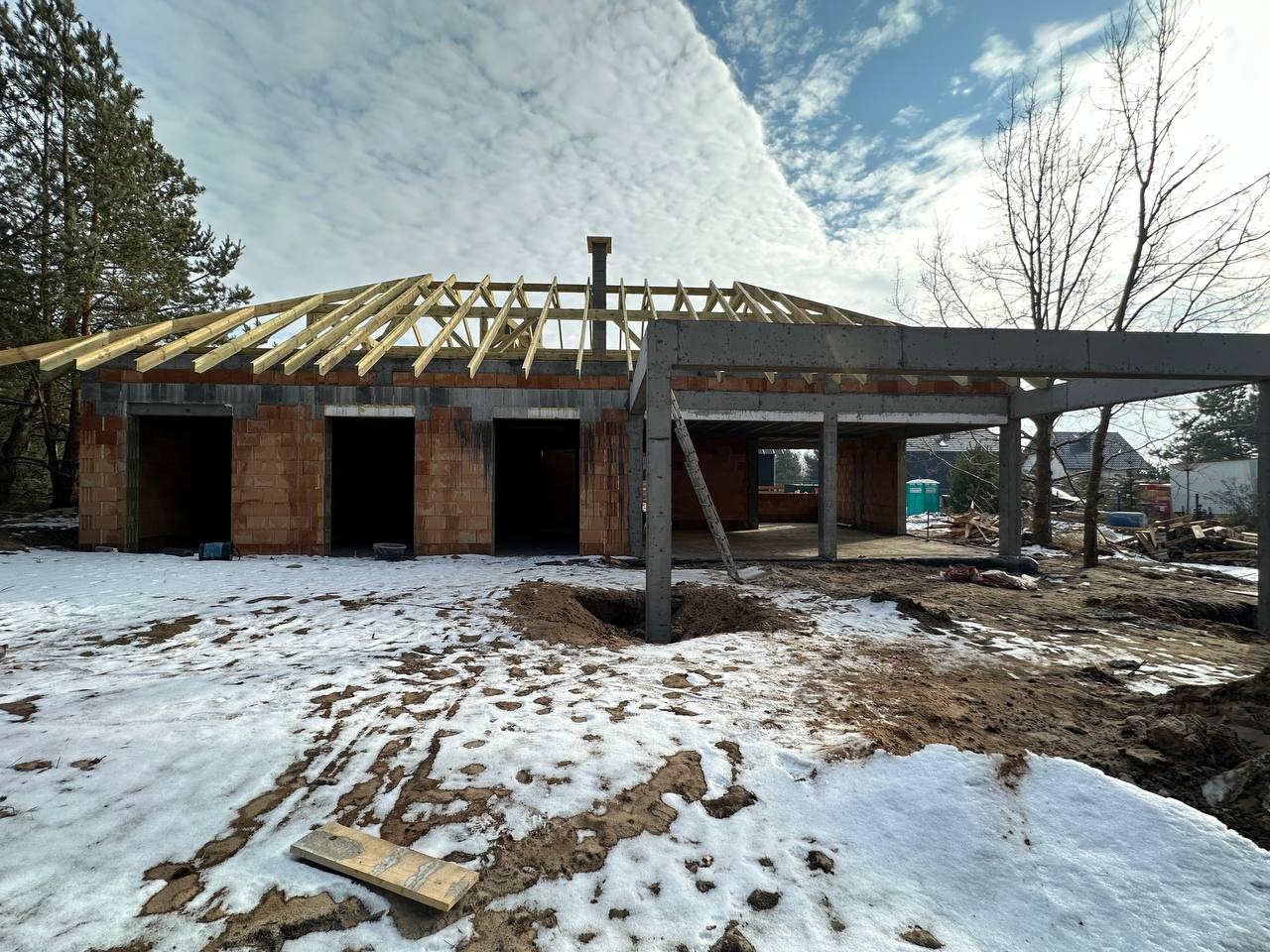 Wznoszony dom murowany z elementami żelbetowymi w zimowej scenerii, widoczne drewniane elementy konstrukcji dachu i częściowo ośnieżony teren budowy.