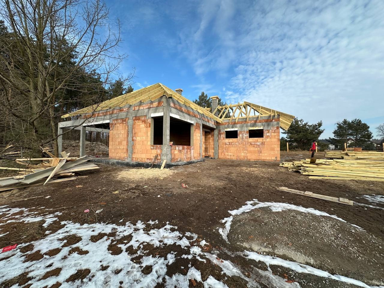 Wznoszenie ścian z czerwonej cegły na parterowym domu jednorodzinnym z widocznymi elementami betonowymi, częściowo odeskowanym dachem i resztkami śniegu na ziemi.