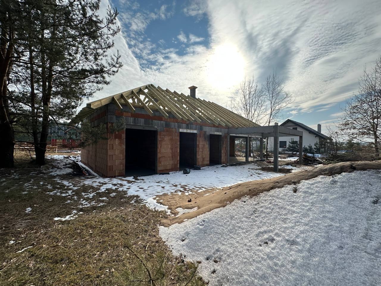 Wznoszenie ścian z czerwonej cegły na budowie domu w zimowej scenerii, widoczne elementy więźby dachowej i zarys przyszłego zadaszenia tarasu.