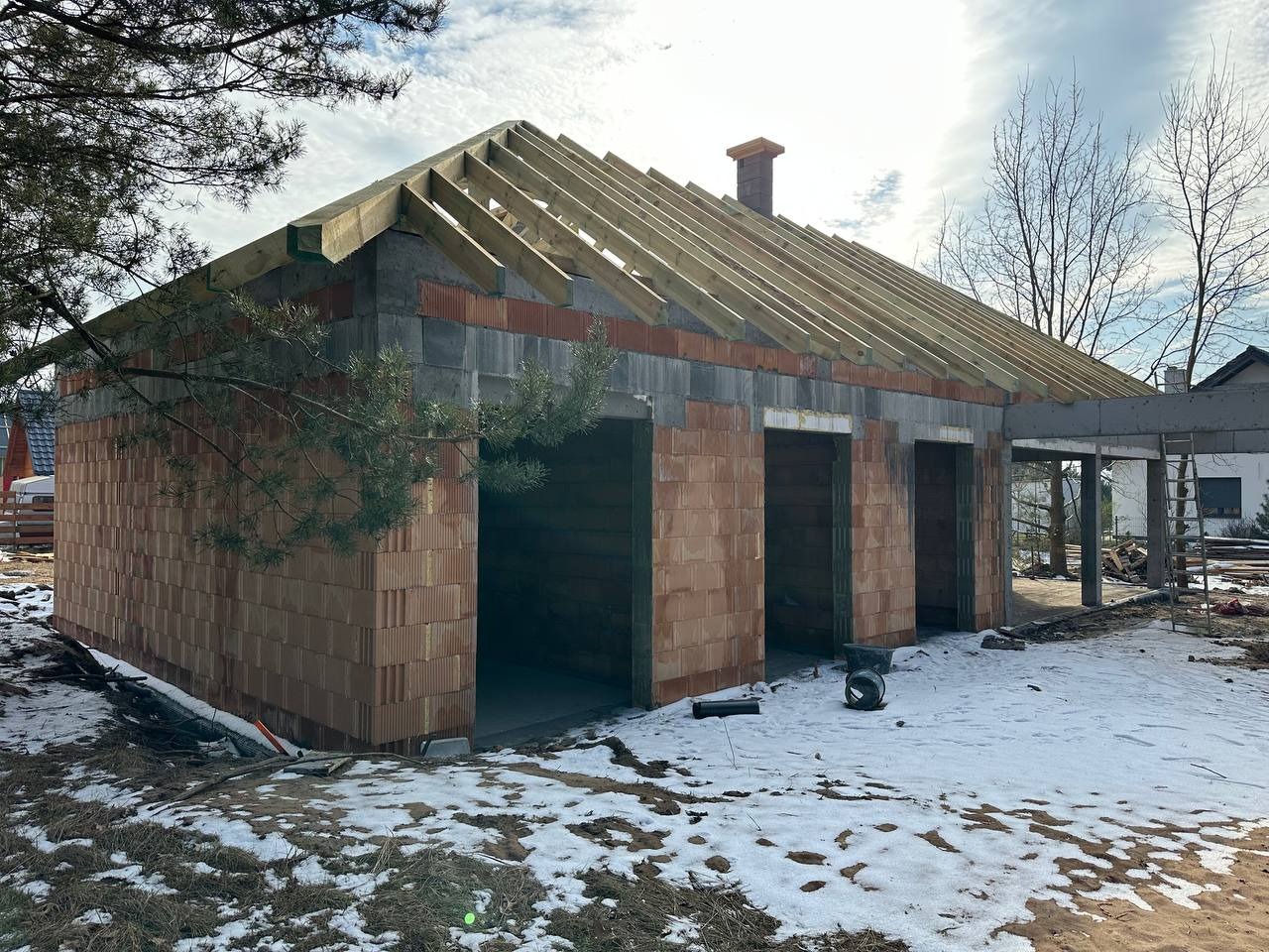 Wznoszona konstrukcja budynku z czerwonej cegły z częściowo wykonanym dachem z drewnianych belek, widok zewnętrzny w zimowej scenerii z resztkami śniegu na ziemi.