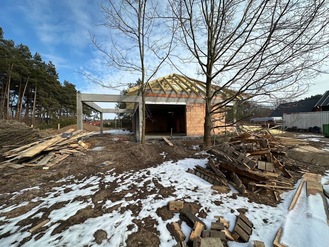 Wczesny etap budowy domu z betonu i cegły, widoczne elementy konstrukcyjne, drewniane szalunki i resztki śniegu na ziemi.