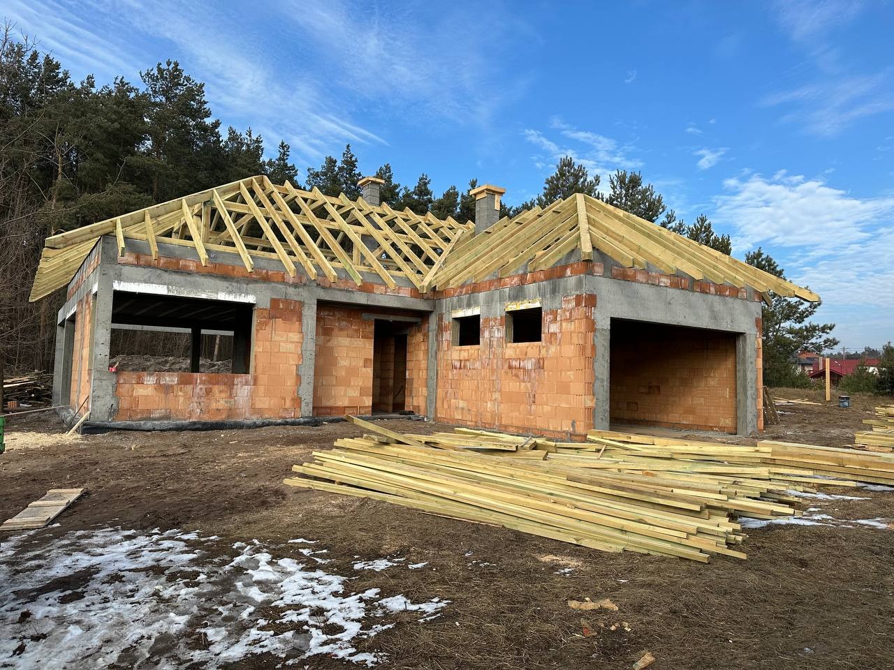 Surowy stan domu jednorodzinnego z czerwonej cegły z drewnianą konstrukcją dachu w trakcie budowy, widoczne kominy i sterta desek na pierwszym planie, las w tle.