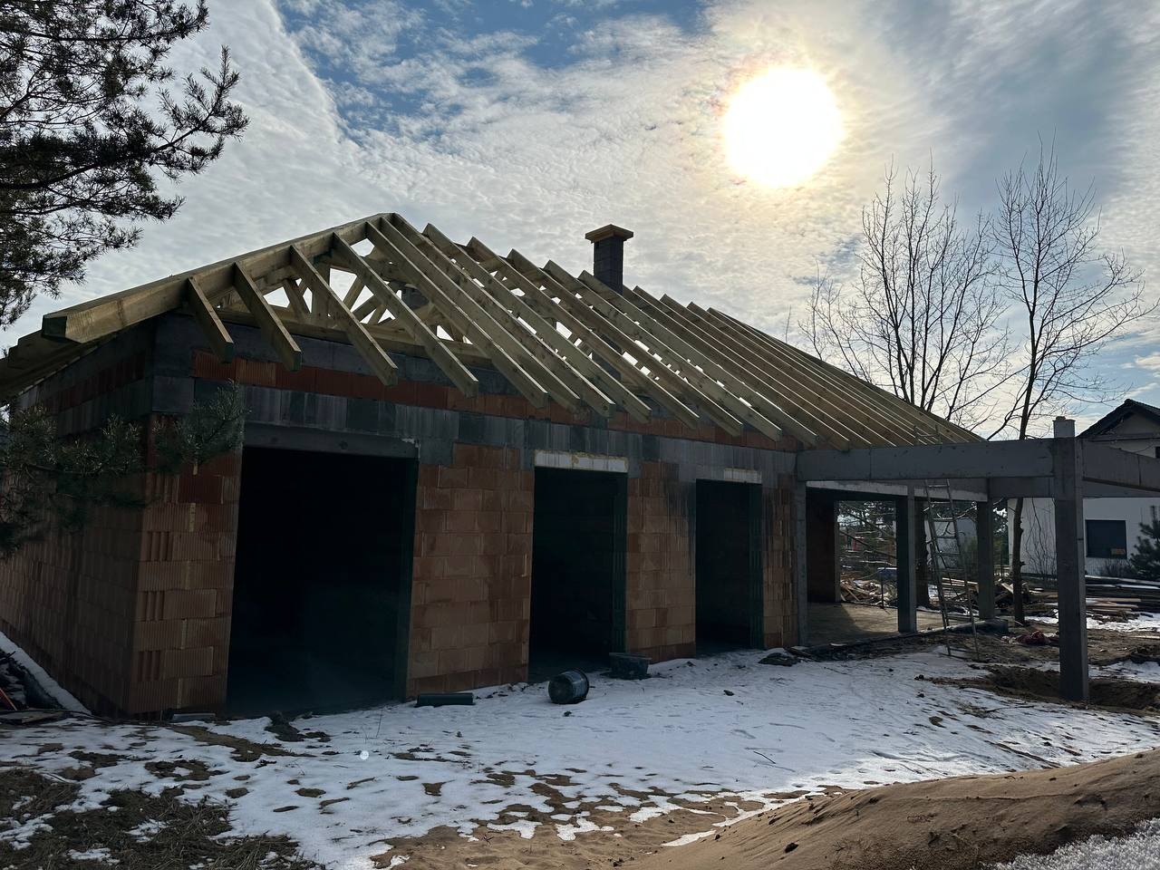 Surowy stan domu murowanego z widoczną konstrukcją dachu z drewna, ściany z czerwonej cegły, częściowo pokryty śniegiem teren budowy i intensywne światło słoneczne w tle.