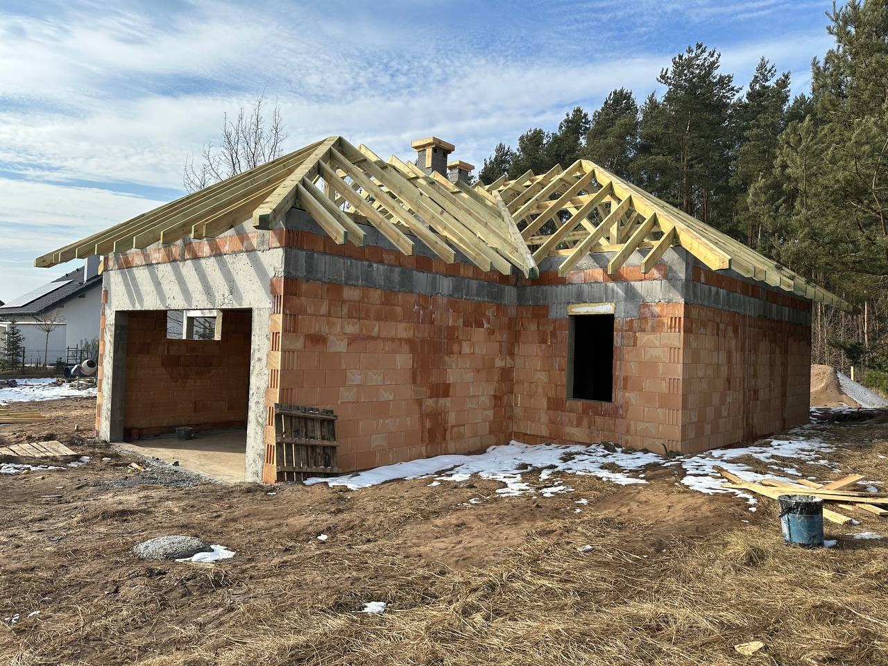 Wznoszenie ścian z czerwonej cegły na parterowym budynku z drewnianą konstrukcją dachu w trakcie budowy, widoczne resztki śniegu na ziemi.