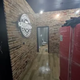 Wnętrze lokalu usługowego z ceglanymi ścianami i podświetlanym logo 'Barber Shop Gentlemen Classic & Modern'. W głębi widoczne drzwi do łazienki z białą toaletą oraz zapakowany towar w folii.